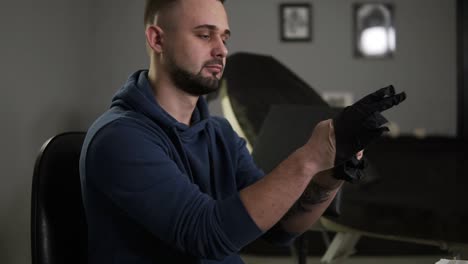 retrato de un artista del tatuaje poniendo los guantes negros de látex antes de una sesión de tatuaje en la tienda de tatuaje. tatuador sentado en una silla en la sala de paredes grises del salón