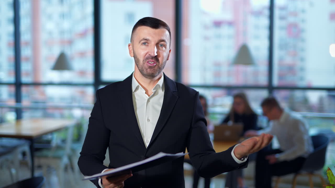 Good-looking energetic bearded man in suit talks to camera holding a folder. Man shows the chart and comments it. Office workers in blur at backdrop.