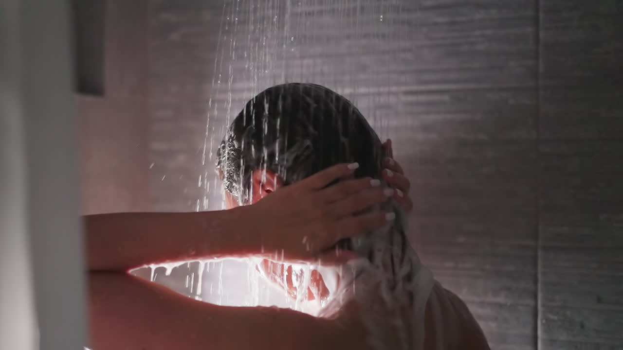 Woman washes hair with shower water closeup. Elegant lady cleans head from foam in bathroom. Woman enjoys routine hygienic procedures at home