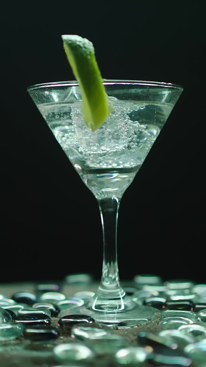 Clear martini glass with lime wedge and ice cubes rotating on black background in elegant composition