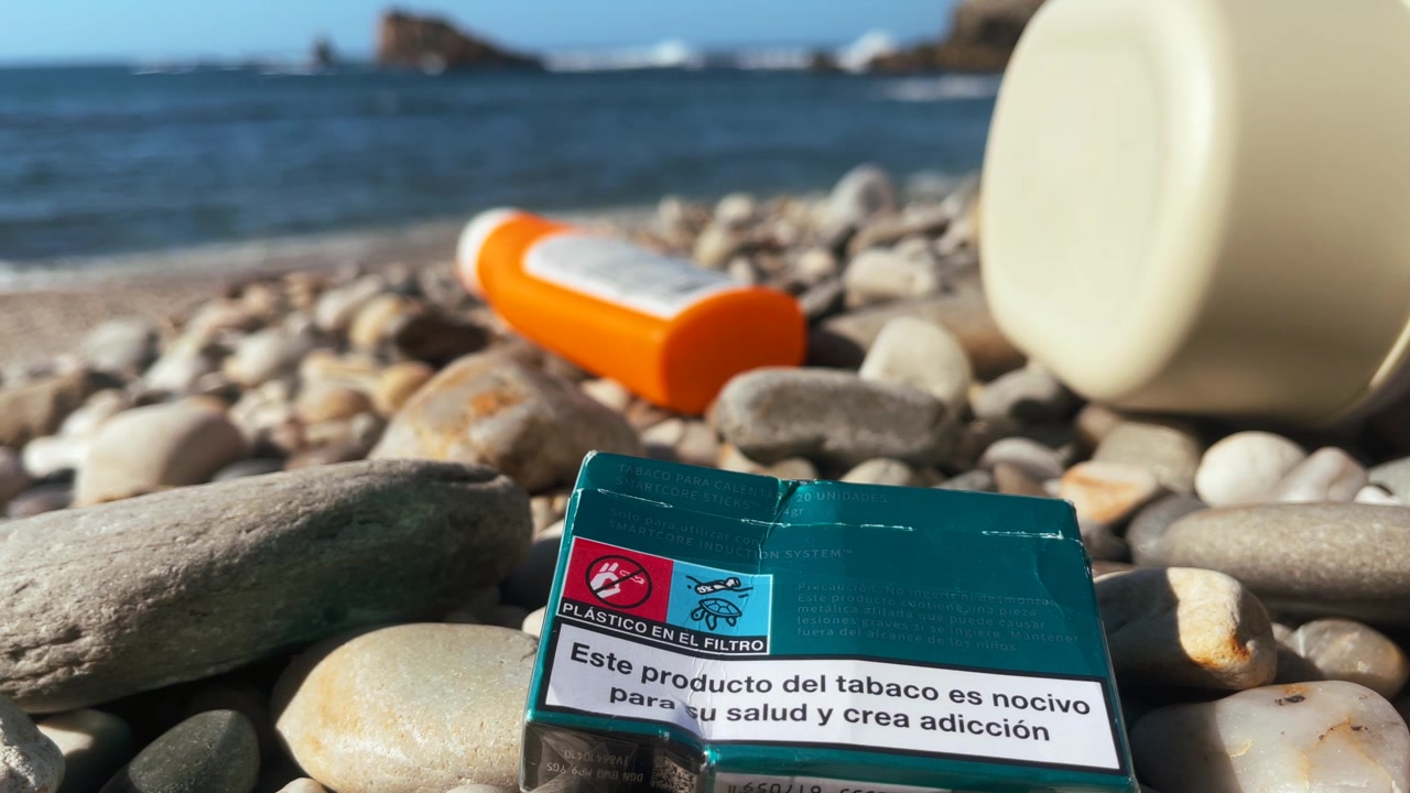 Discarded cigarette box and plastic waste on rocky beach shore, symbol of pollution