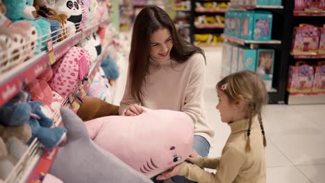 Mother shows big soft toy to her daughter in children&#039;s department store, both smiling