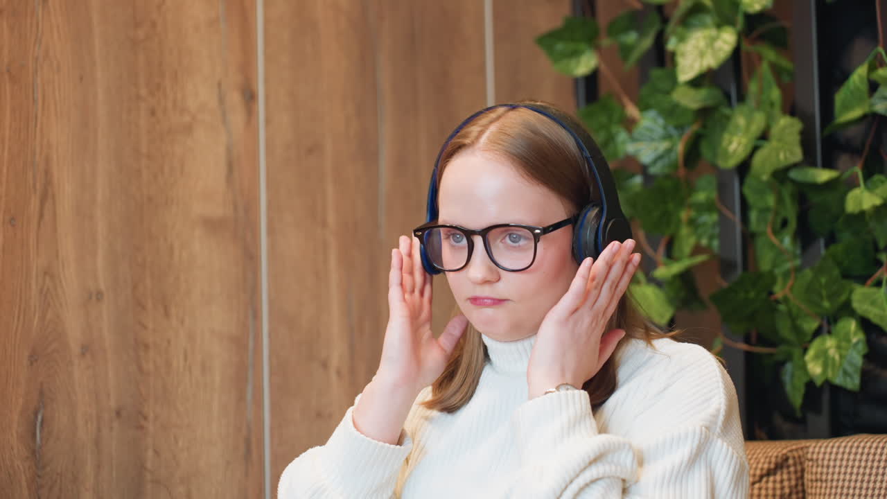 Girl in white sweater and glasses lifts headphone toward her head with playful expression, preparing to enjoy music and dance in cozy indoor setting with wooden walls and green plants