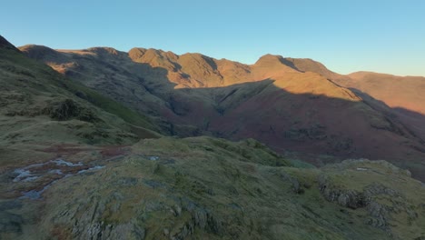 Flying over shadowed crags towards dawn lit mountaintops with reveal of deep shadowed valley. Langdales, English Lake District, Cumbria, UK.