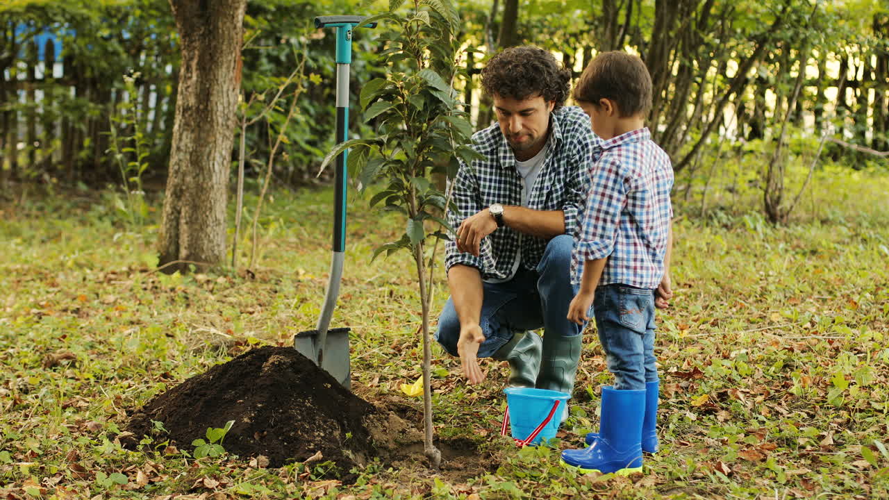 retrato de un niño y su padre plantando un árbol. el padre le explica algo a su hijo y pone la tierra con la pala de juguete en el cubo de juguete. hablan y sonríen. fondo borroso