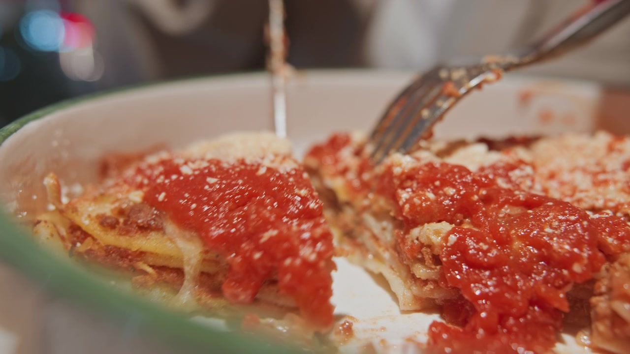 Primo piano di una persona che taglia la lasagna con salsa di pomodoro rossa e formaggio fuso in un piatto bianco. Le forchette vengono usate per separare le fette, rivelando gli strati all'interno del pasto di pasta al forno. Illuminazione soffusa.