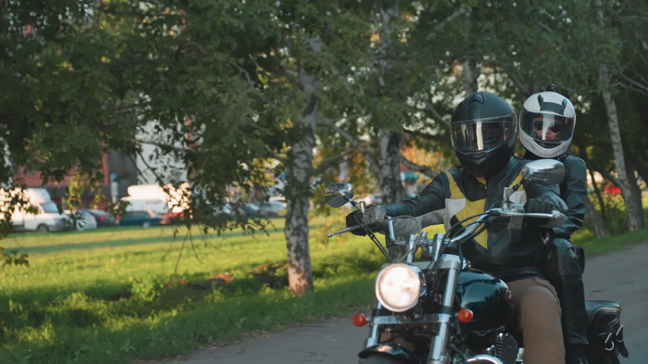Motorcyclist with passenger rides along city road past tree lined sidewalk and residential buildings, motorcycle headlight shining while cars and urban background