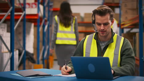 male worker wearing headset sitting at desk working on laptop in busy warehouse