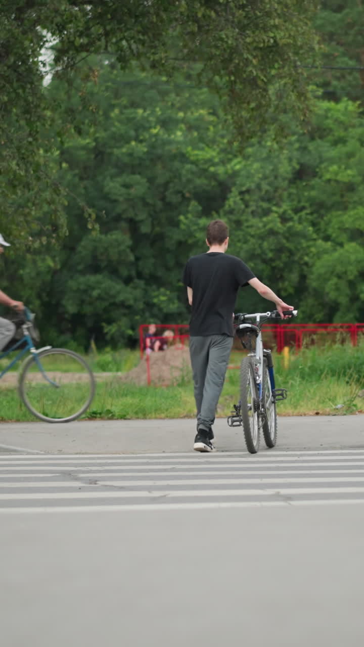 두 형제가 자전거를 타고 보행자 통로를 가로질러 나란히 걸어가는 뒷면, 배경에는 흐릿한 사람들이 고, 초록색 나무, 밝은 빨간색 울타리가 있습니다.