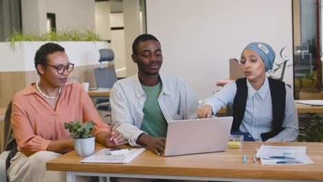 young worker working with laptop sitting at his desk while muslim businesswoman and businesswoman talk to him sitting at desk