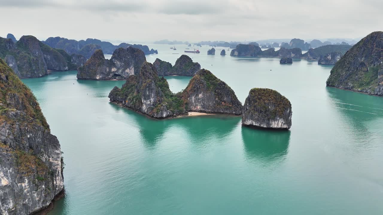 Turquoise waters surround towering limestone islands in Ha Long Bay, Vietnam, with scattered beaches and distant boats visible beneath a cloudy sky, creating a serene coastal seascape view