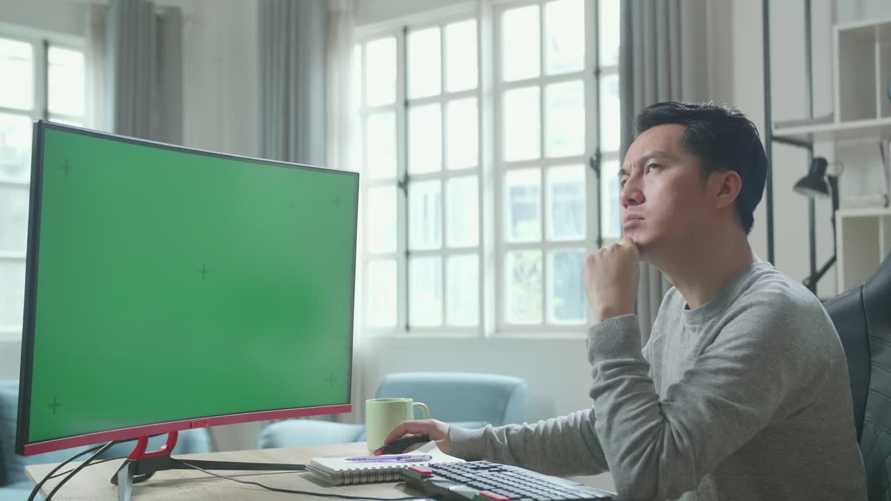 Asian Man In Long Sleeved T-Shirt Thinking Then Typing On Green Screen Desktop Computer While Working At Home.