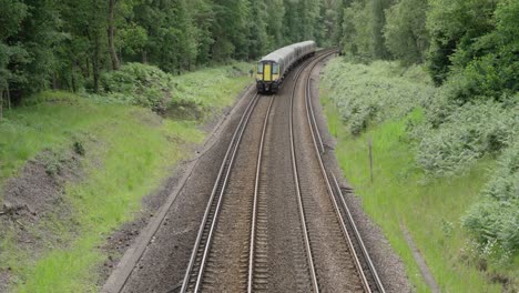 a modern british commuter train departs through green countryside on a curved double-track railway, surrounded by summer foliage and forest under an overcast sky.