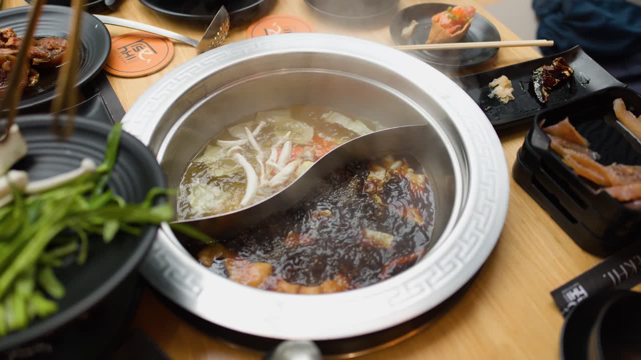 Hands add vegetables and sliced meat to steaming dual-broth hotpot under bright restaurant lighting