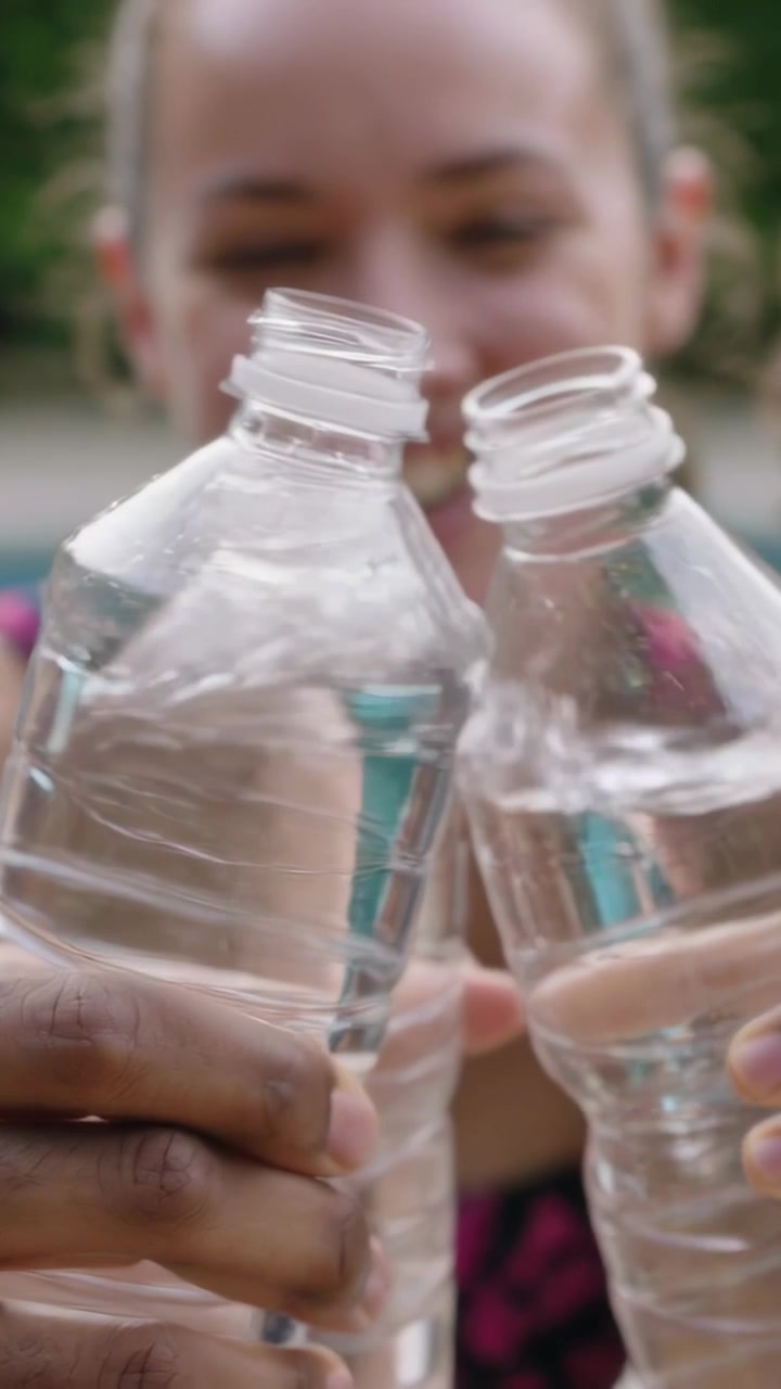 Vidéo verticale : La caméra recule, une main et une femme trinquent avec des bouteilles d'eau, simulant un toast au bord de la piscine, en haut de maillot de bain.