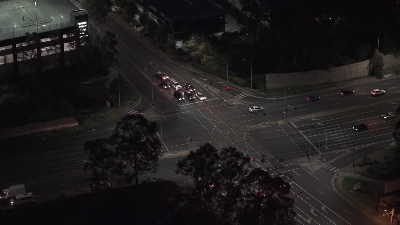 Aerial footage captures a bustling nighttime intersection in Australia, showcasing city lights and traffic flow. The overhead view highlights urban planning and street design