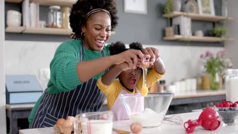 happy african american mother and daughter adding egg to bowl in kitchen, slow motion