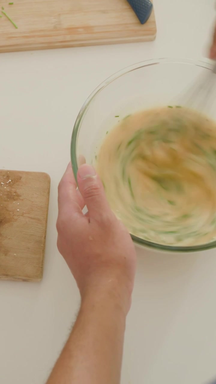 Vertical video: Steadying bowl, cook whisking egg-herb mix with whisk in kitchen for batter