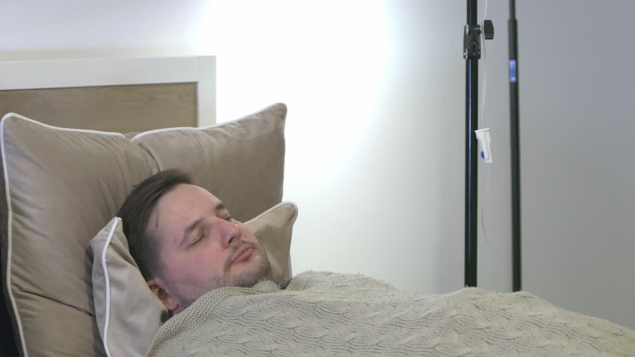 paciente varón cansado durmiendo en una sala privada con goteo intravenoso balanceándose suavemente al lado de la cama