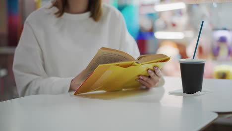 close-up of hands flipping through pages of a yellow book on a table with coffee cup nearby, soft lighting and blurred background featuring colorful toys and vibrant decor in a relaxed mall setting