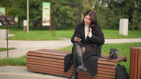 woman seated with crossed legs holding phone, adjusting her hair in relaxed park setting, surrounded by greenery, wearing black coat and boots, with bag placed beside her on wooden bench