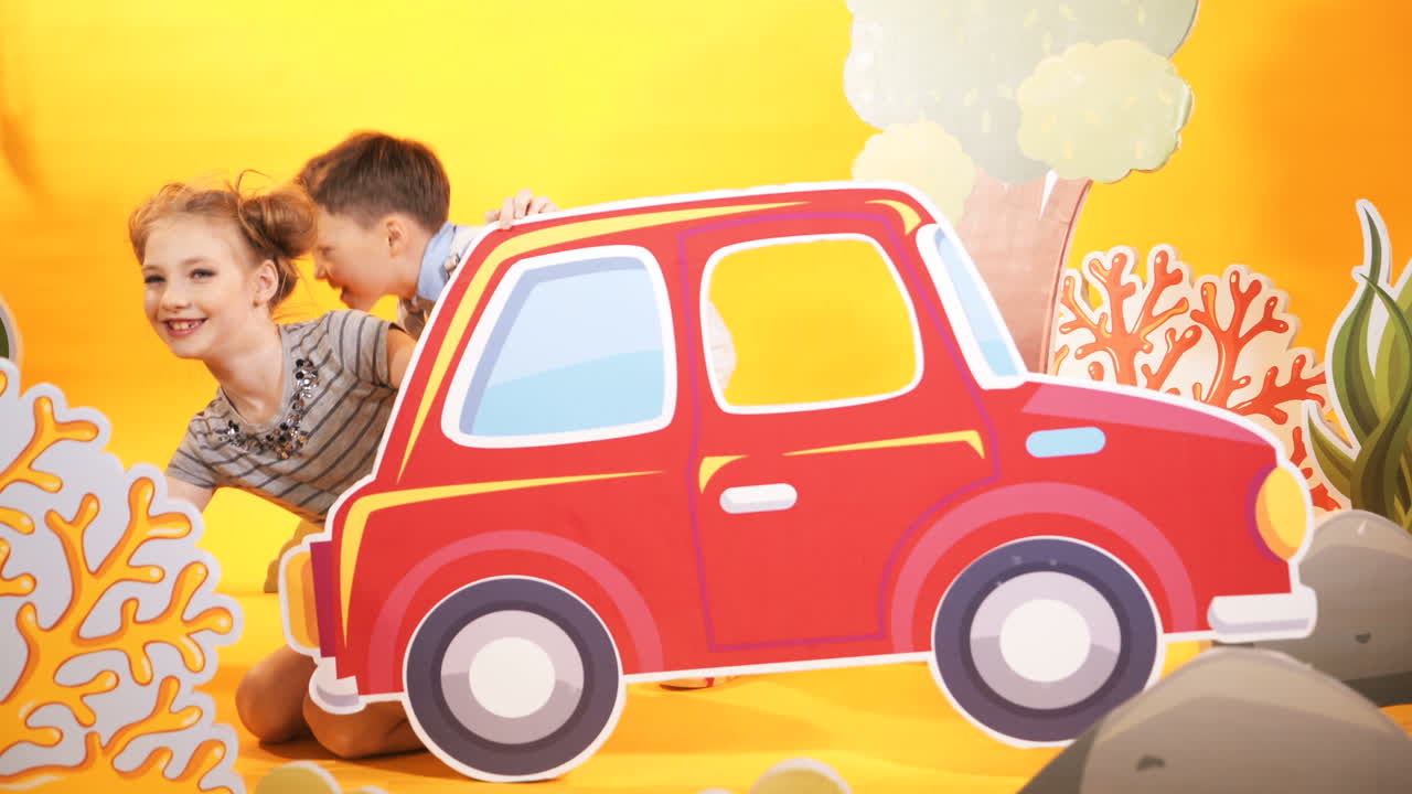 Happy boy and girl playing together in the children's studio room with decor over the yellow background. Beautiful toyful carton scenery of a big red car and colorful bushes.