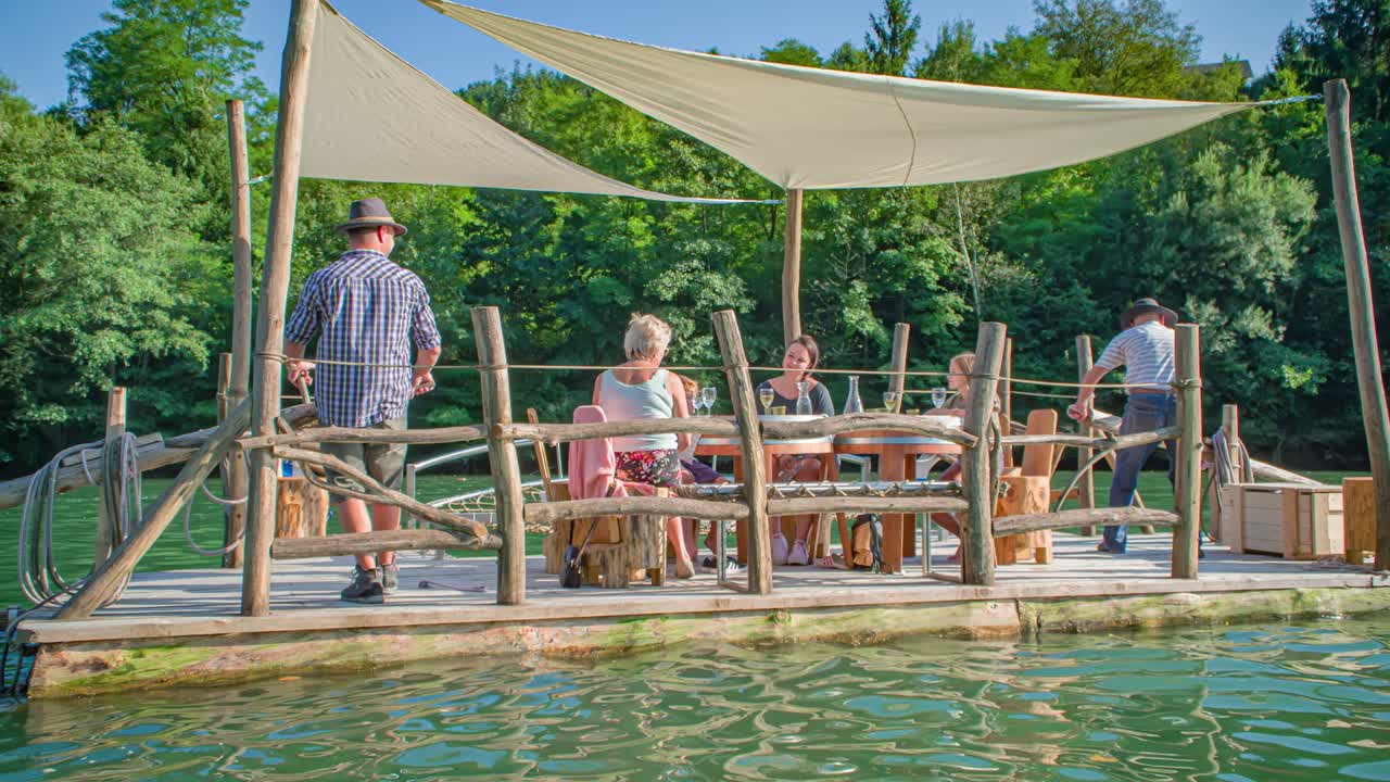Family takes a tour and picnicking on a large raft with chairs on calm water in a colorful landscape on a sunny day. Tracking shot