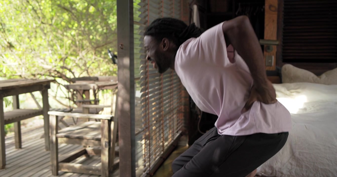 Dreadlocked African American man in pink tee rubbing lower back on bed relieving pain, copy space