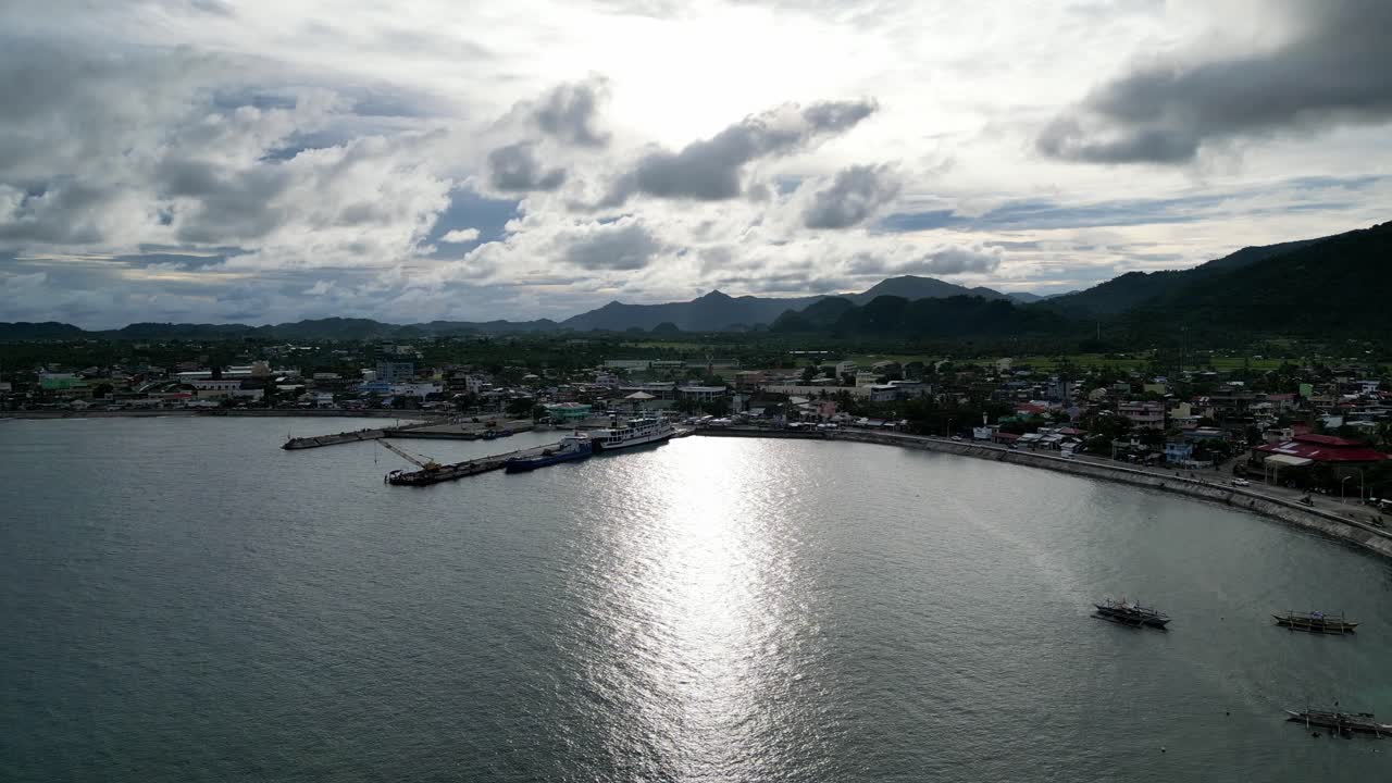 Cinematic, Establishing Aerial View of wide ocean bay alongside idyllic island town of Virac, Catanduanes, with bangka boats, simple homes and dramatic clouds