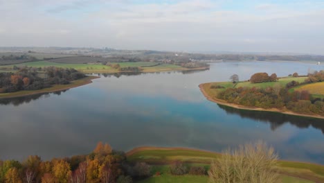 Forward-movement-drone-4k-over-reservoir-in-England-in-autumn-with-glassy-reflection-of-the-sky,-fishing-boats-and-natural-forest