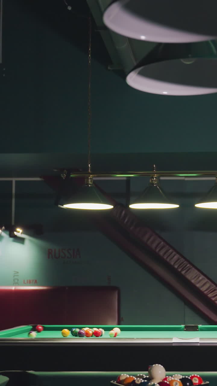 Giocatore di biliardo in camicia bianca si china su un tavolo da biliardo verde, concentrandosi intensamente sul tiro alla palla bianca. La sala da gioco con luci soffuse e pareti scure crea un'atmosfera rilassata e informale