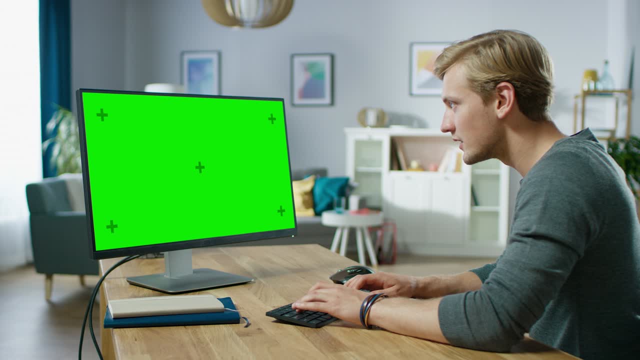Giovane uomo attraente lavora su un computer personale con schermo mock-up verde, seduto alla sua scrivania nell'accogliente appartamento