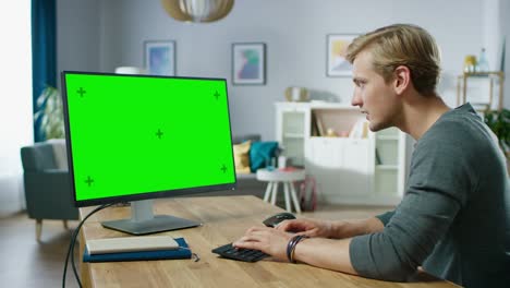 un joven guapo trabaja en una pantalla de maqueta verde de una computadora personal mientras está sentado en su escritorio en el acogedor apartamento.