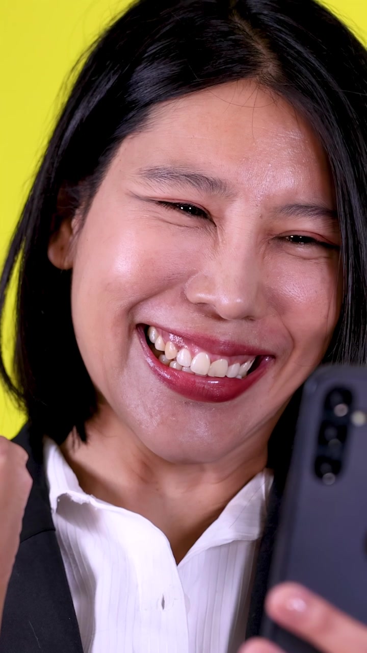 Close-up businesswoman reacts with joyful surprise to smartphone message, bright yellow studio background, static framing