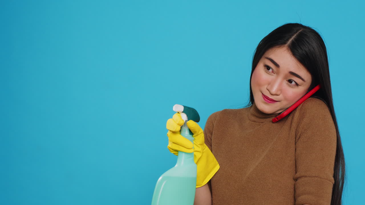 Asian housekeeper using chemical detergent and duster to clean house while talking at mobile phone