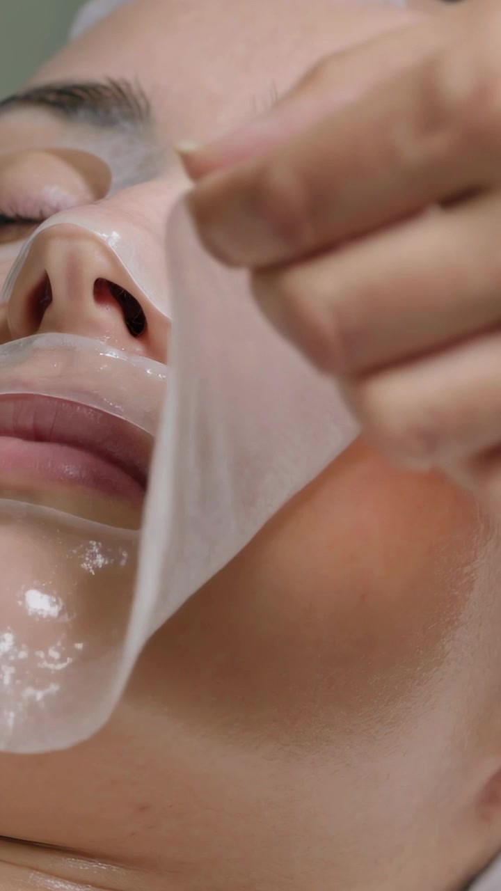 Vertikal video: En hånd kommer inn og skreller av en hydrogelmaske fra en kvinne på en spabenk, avslører duggfrisk hud.