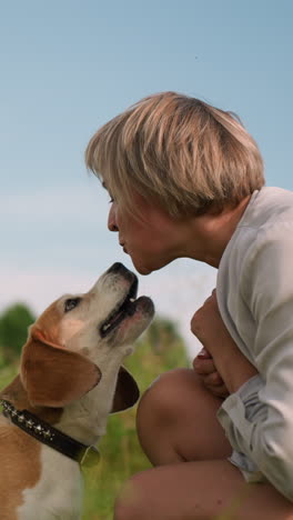 dog owner interacting playfully with her pet as they share a tender moment in outdoor grassy field, owner is squatting down on sunny day while the dog jumps to kiss her