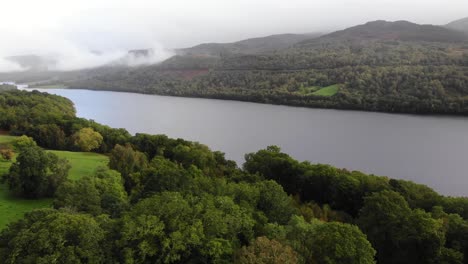 Vista-Aérea-De-Los-árboles-De-La-Orilla-Del-Río-Del-Valle-Verde-Con-El-Lago-Tummel-En-El-Fondo