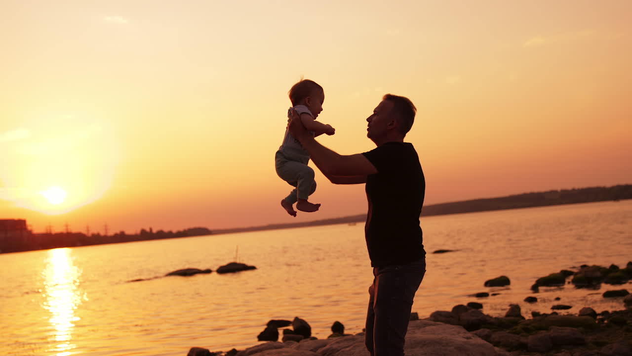 Father and baby boy spending time at the river bank. Dad tossing up his little son at backdrop of orange sky at sunset.