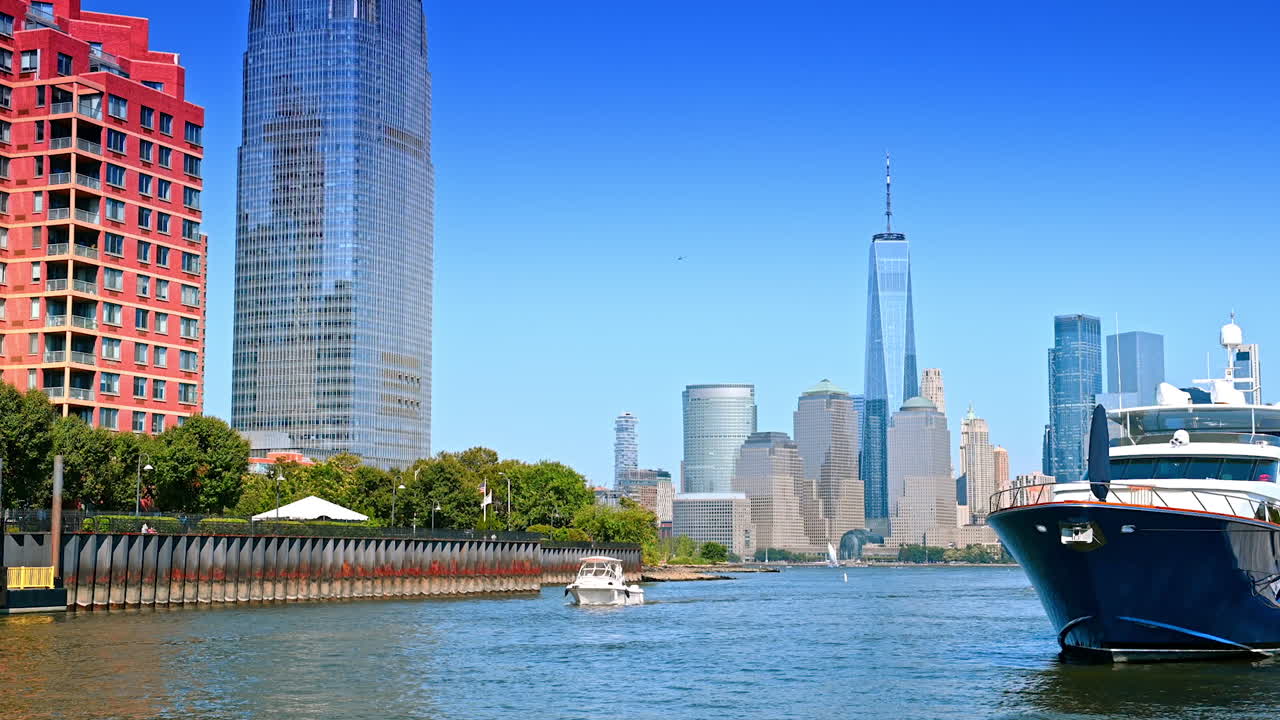 Little white boat approaches the camera along the green waterfront. Large cruise boat stands nearby on the anchor. Skyline of Manhattan at backdrop