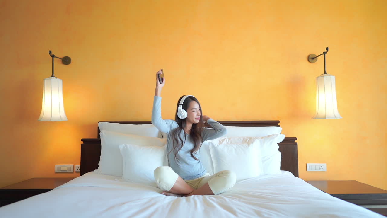 Wide shot of young pretty Asian woman sitting on a hotel bed listens to music through her headphones while she dances to the music. copy space