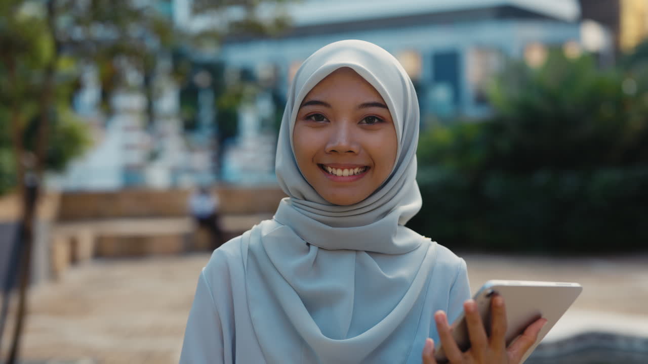 Portrait of a smiling young woman in a hijab holding a tablet outdoors