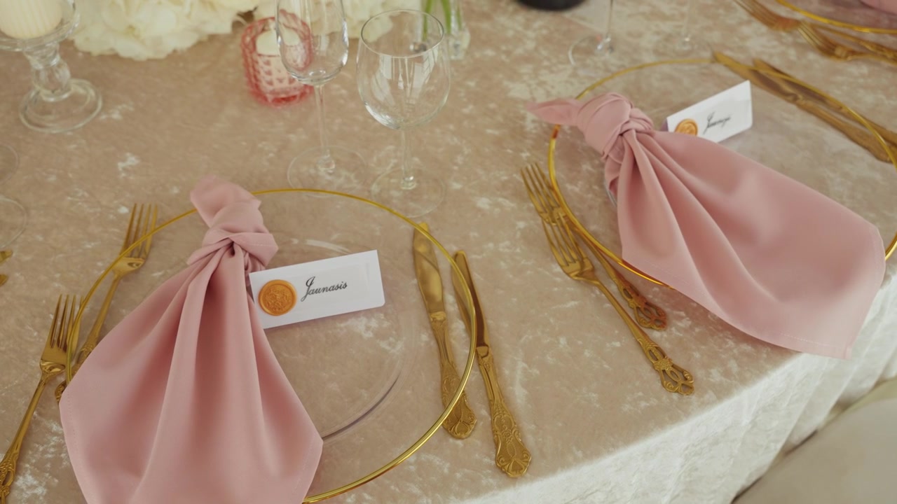 Close-up shot of a luxurious wedding table elegantly set with gold cutlery, blush pink knotted napkins, glass plates, and personalized name cards, under soft natural lighting.