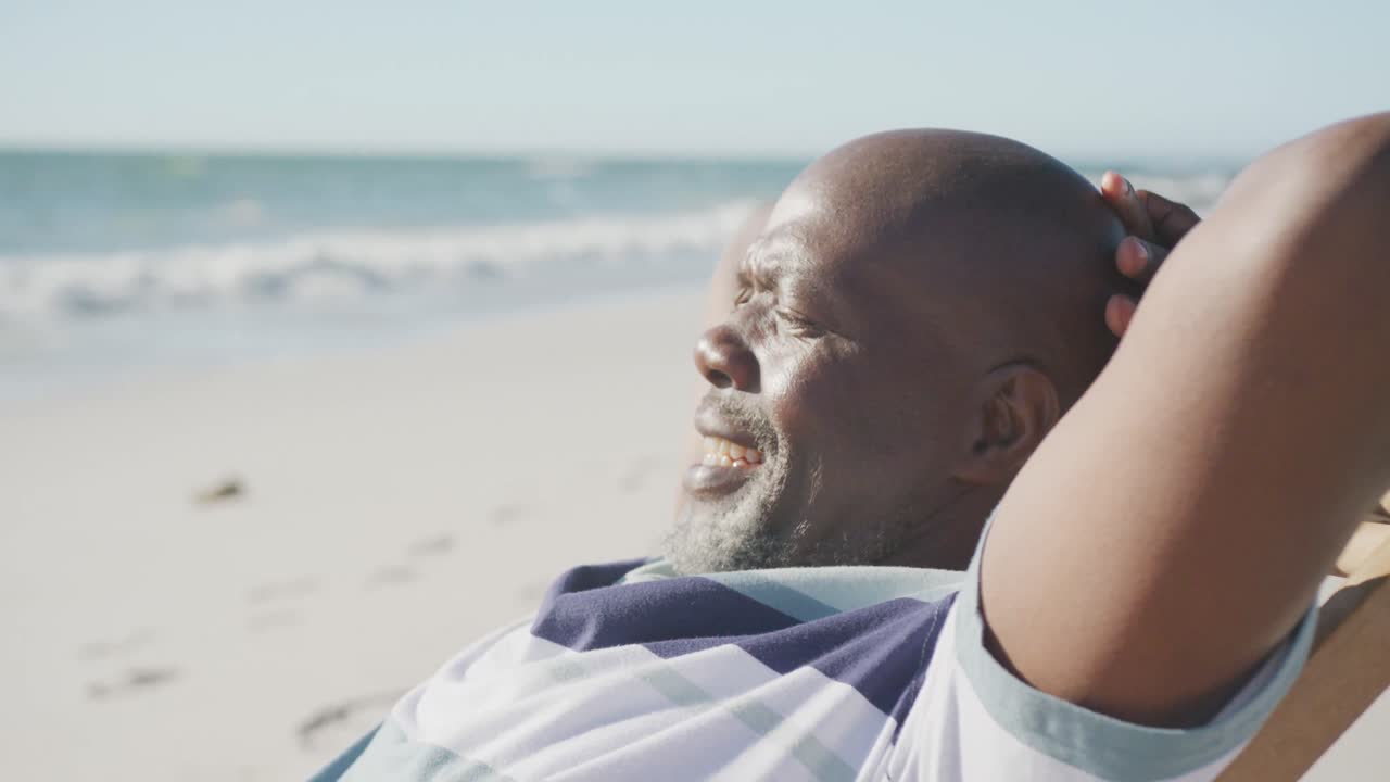 Happy senior african american man sitting on deck chair at beach, in slow motion
