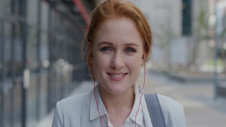 ritratto attraente giovane donna d'affari con la testa rossa sorridente che indossa auricolari che si gode di ascoltare musica in slow motion in città urbana