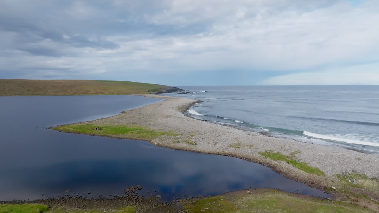 Sweeping aerial view of a coastal lagoon and pebble beach on the Isle of Lewis in the Outer Hebrides, Scotland. Calm Atlantic waves meet rugged terrain under moody skies — a wild island landscape