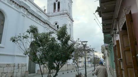 Increíble-Campanario-Blanco-De-La-Iglesia-En-Un-Día-Nublado-Inclinándose-Hacia-Abajo-Para-Revelar-Ancianos-Caminando-Por-La-Calle-Ecuador