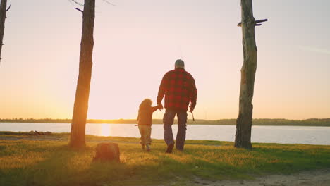 old man and his grandson are travelling in nature walking to lake for fishing in sunset rear view