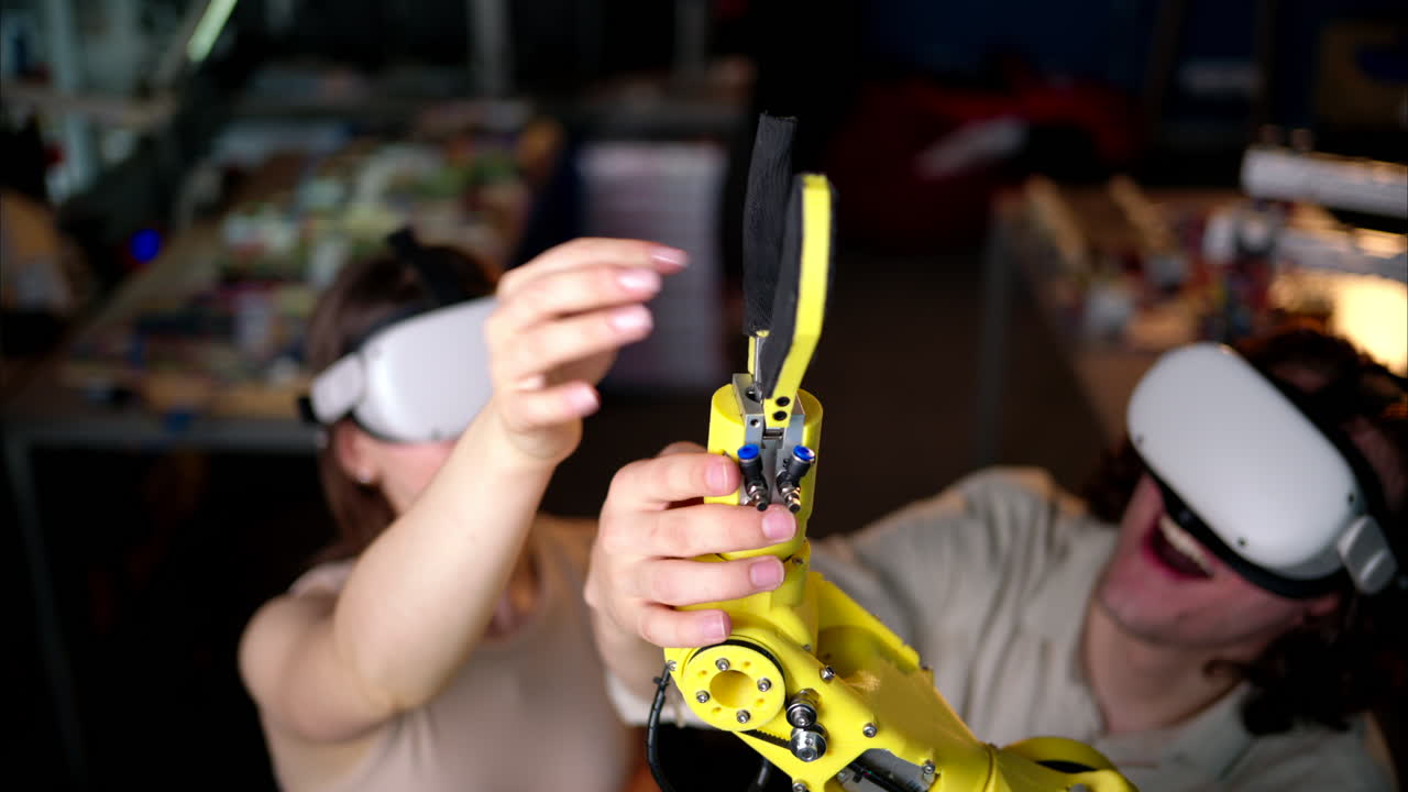 Two young happy engineers with VR virtual reality headsets fixing an yellow robotic arm in the workshop, computer programming, celebrating success