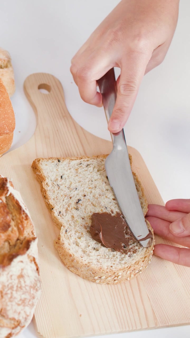 Close-up of hands spreading chocolate hazelnut cream on bread over a wooden board.
